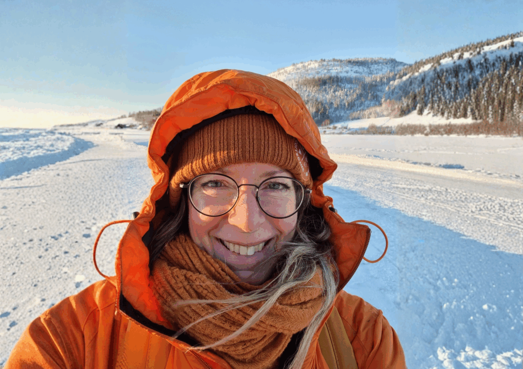 Kvinna stående i ett snölandskap iklädd en orange vinterjacka leende mot kameran.