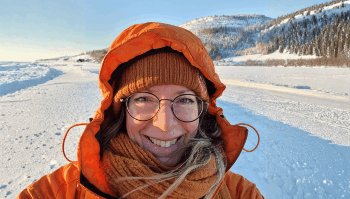 Kvinna stående i ett snölandskap iklädd en orange vinterjacka leende mot kameran.